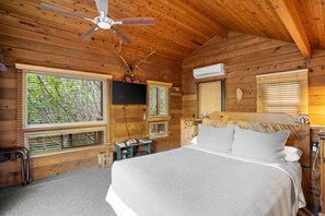 Une chambre dans une cabane en bois, équipée d’un lit, d’un ventilateur de plafond, d’un appareil de climatisation et de fenêtres avec des stores.