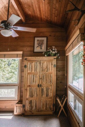A wooden cabin interior with a ceiling fan, a wooden wardrobe, and large windows.