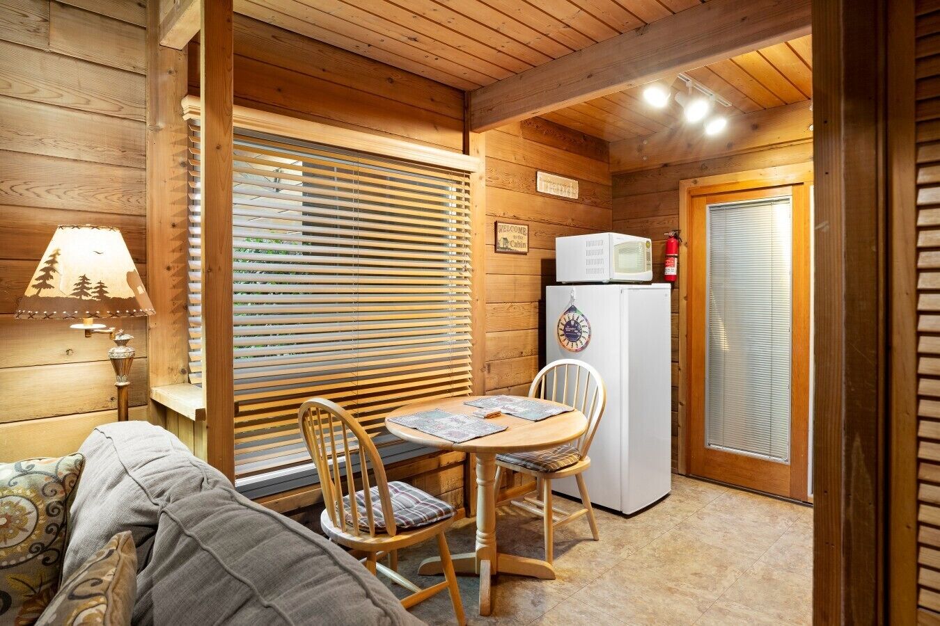 A cabin interior with a wooden ceiling, a table with chairs, a refrigerator, and a lamp.