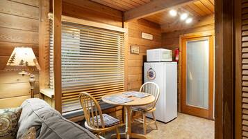 A cabin interior with a wooden ceiling, a table with chairs, a refrigerator, and a lamp.
