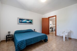 Een slaapkamer met een bed, een nachtkastje, een stoel, een deur naar een eetgedeelte, en een abstract schilderij aan de muur.
