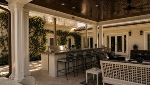 An outdoor patio with a bar, seating area, and a view of a building with columns and greenery. - Pelican Beach Mansion (Fort Lauderdale)