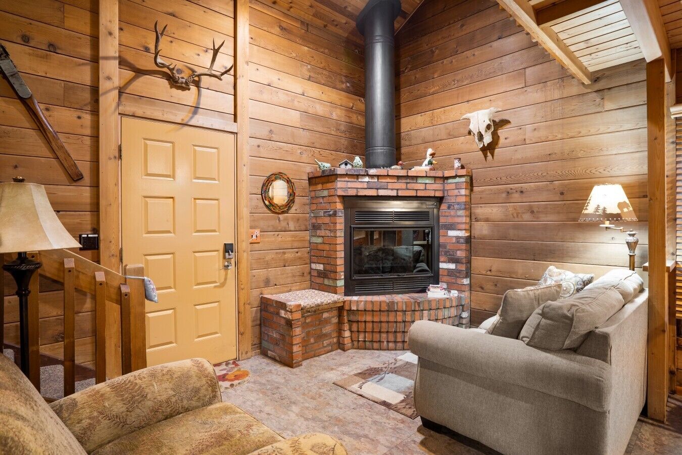 A cozy cabin interior with a brick fireplace, wooden walls, a door, and a sofa.