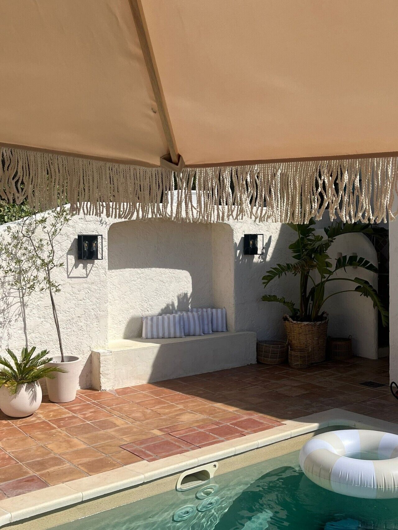 Zona de piscina con un flotador blanco, un banco con cojines y plantas en macetas.
