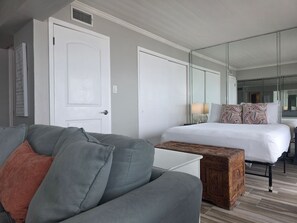 Ein modernes Hotelzimmer mit grauem Sofa, einer Holzablage, einem Bett mit gemusterten Kissen und einem großen Spiegel.