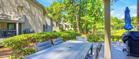 A covered outdoor patio with a table and chairs, surrounded by greenery and a view of a residential area.