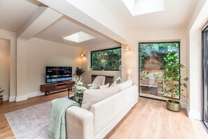 A modern living room with a sofa, TV, and a large window with a view of greenery. - Luxury 2BR Apartment, Garden & Central Access - Pass the Keys (London)