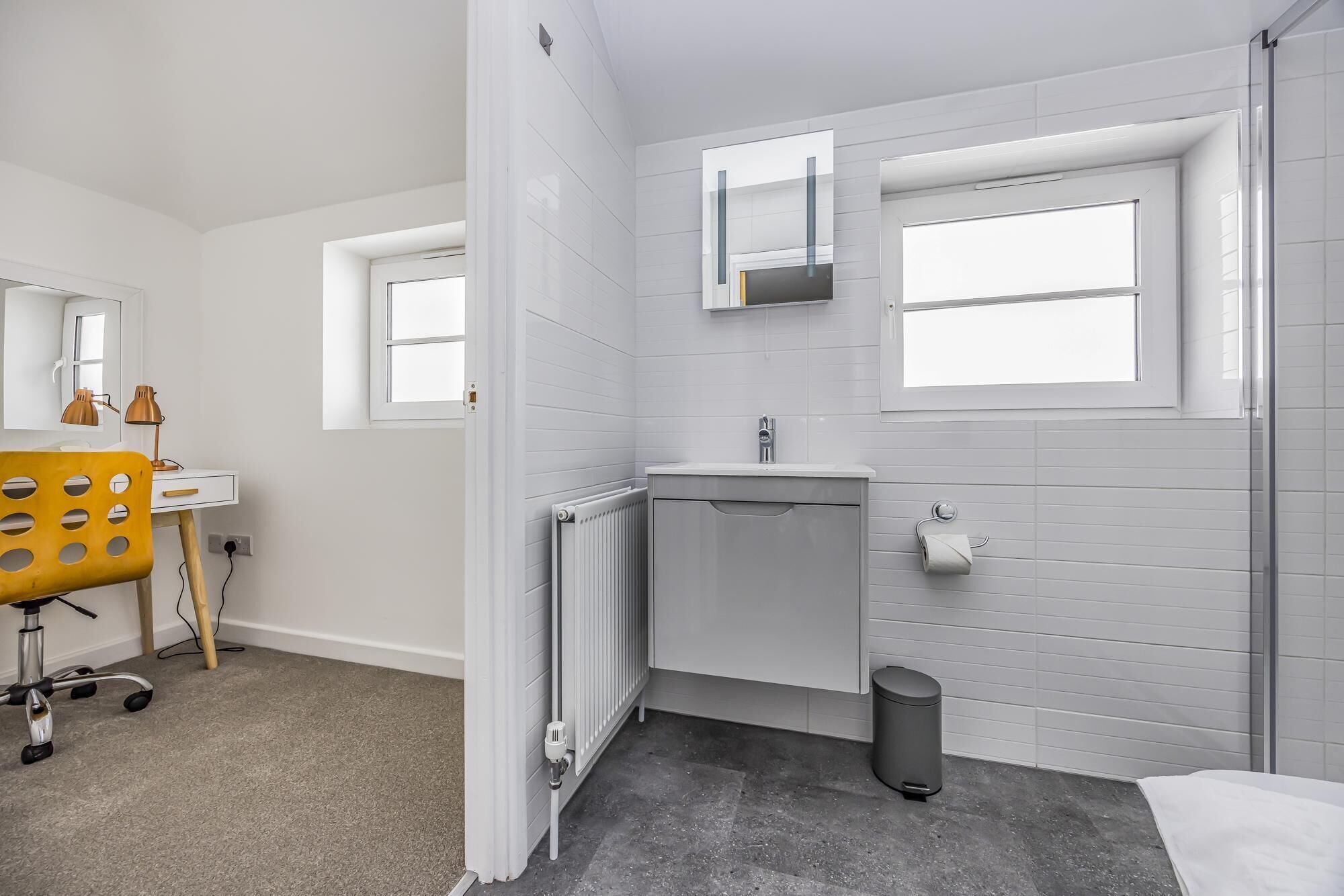 Une salle de bain avec des murs recouverts de carreaux blancs, un lavabo blanc, un miroir au-dessus du lavabo, un radiateur, une poubelle et une chaise jaune.