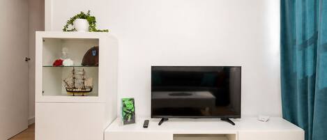 A modern living room with a flat-screen TV on a white entertainment unit, a small shelf with decorative items, and a door leading to another room.