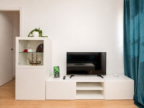 Ein modernes Wohnzimmer mit einem Flachbildfernseher auf einer weißen Unterhaltungseinheit, einer kleinen Ablage mit Dekoration und einer Tür zu einem weiteren Raum.