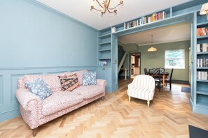 Ein Zimmer mit einer gemusterten Couch, einem gestreiften Sessel, einem Holzboden im Fischgrätmuster und einem Bücherregal voller Bücher.