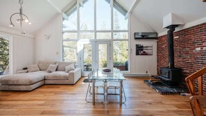Een ruime kamer met een groot raam, een houten vloer, een glazen tafel met stoelen, een bank en een open haard.