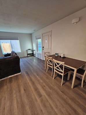 Ein geräumiger Wohnbereich mit einem dunkelgrauen Sofa, einem Holzespeisetisch mit vier Stühlen und einem großen Fenster, das für natürliches Licht sorgt.