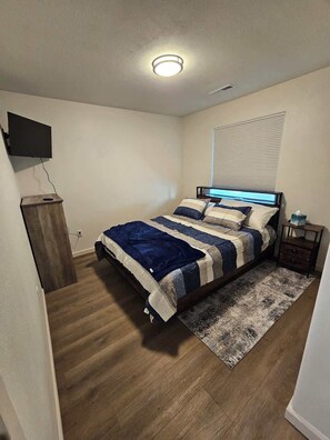 Ein Schlafzimmer mit Bett, einem an der Wand montierten Fernseher, einem Nachttisch und einem Fenster mit Jalousien.