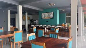 Un grand espace dédié à la salle à manger, avec des tables en bois et des chaises à l’assise bleue, un coin cuisine aménagé avec un buffet, et un mur d’accent de couleur turquoise orné d’un tableau représentant un paysage.