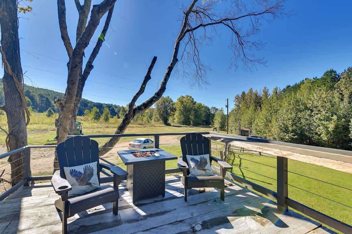 A deck with Adirondack chairs and a table, overlooking a road and greenery.