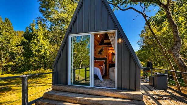 A cabin with a glass door entrance, a wooden deck, and a view of the surrounding forest. - Eagle’s Nest Couples Escape w/ Hot Tub & Firepit (Cherokee)