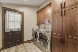 A laundry room with two front-loading washing machines, wooden cabinets, and a door with a window. - Luna’s Mountain Manor | Chef Kitchen & Game Room (Stateline)