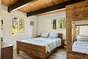 Un dormitorio con cama de madera, dos ventanas, una mesita de noche y una estructura de techo de madera.