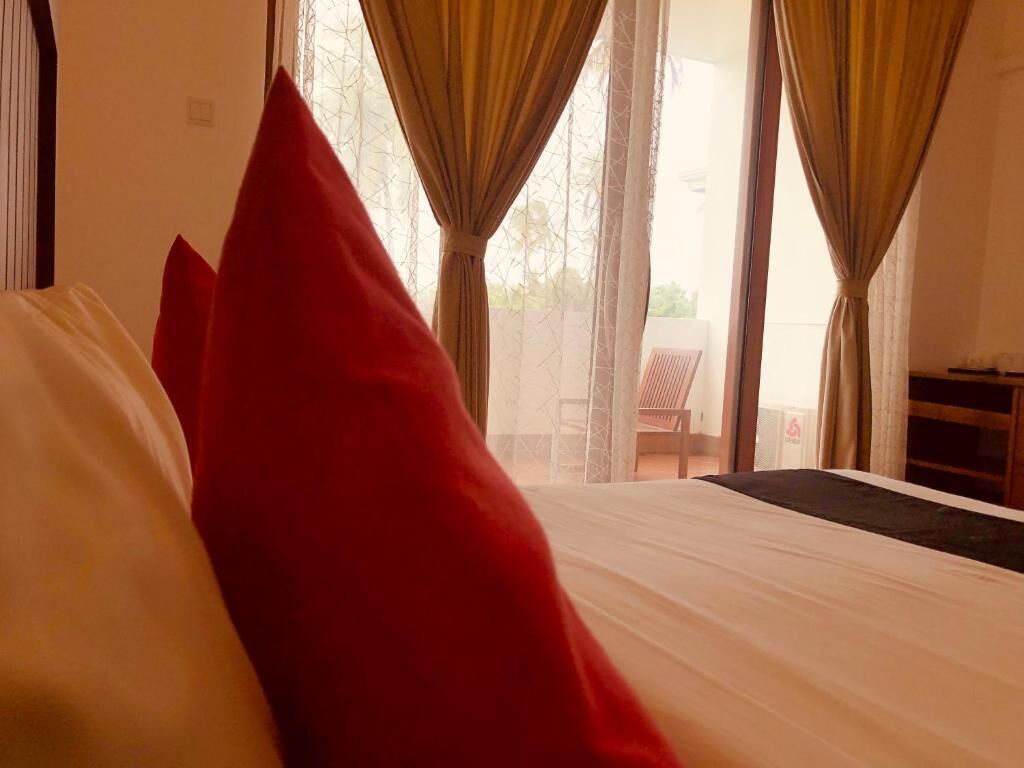 A bed with a white sheet and a red pillow, a window with curtains, and a view of the outside.