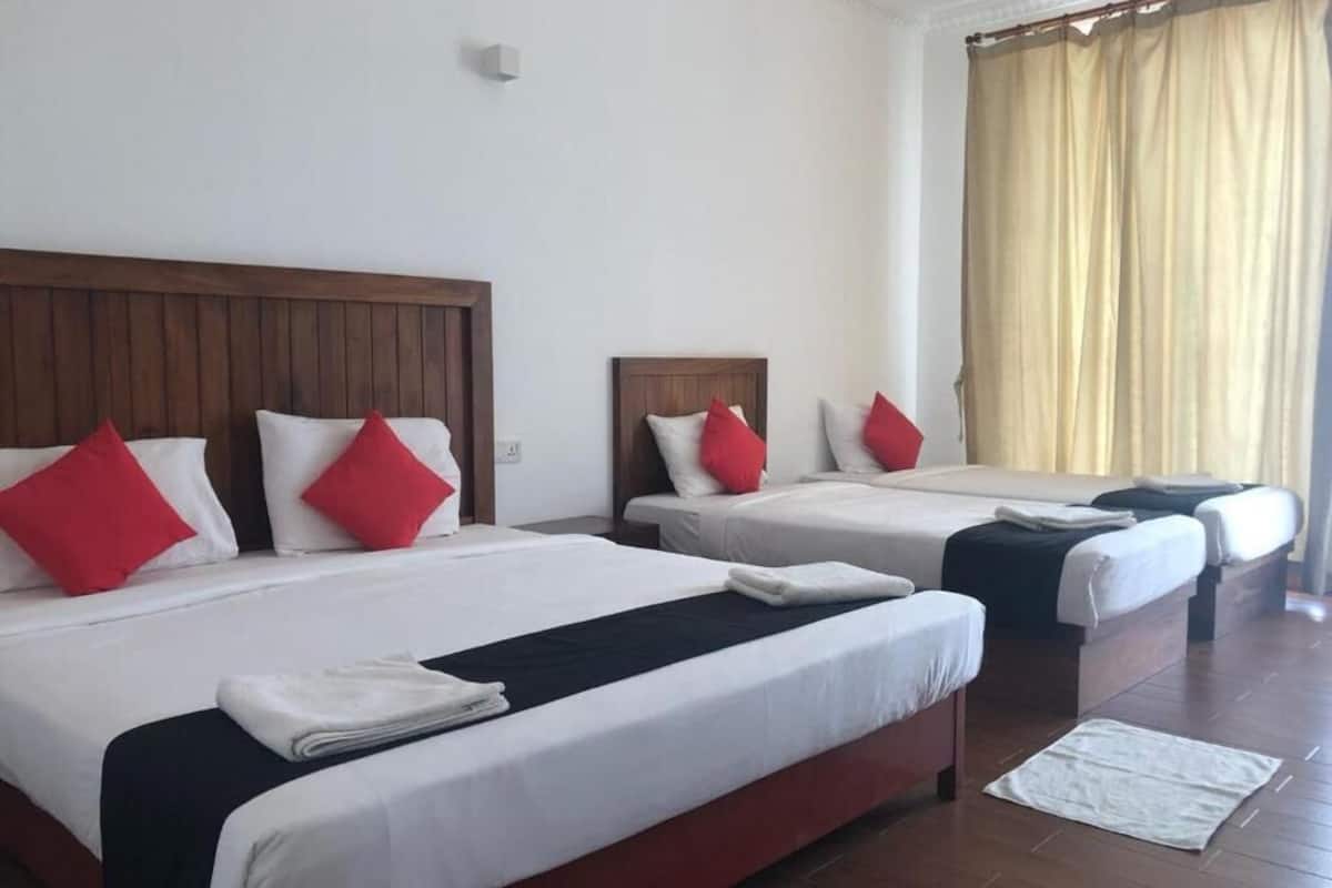 A hotel room with three beds, each with white bedding and red pillows, wooden headboards, and a large window with beige curtains.