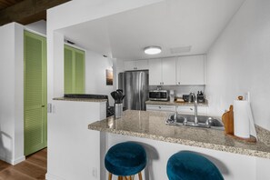 A modern kitchen with white cabinets, a stainless steel refrigerator, a microwave, and a sink. There are two blue bar stools and a green door. - Kona Mansions#226 (Kailua-Kona)
