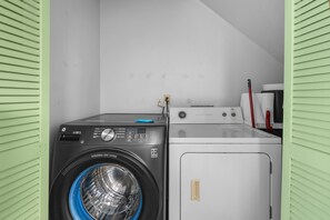 A laundry room with a front-loading washer and dryer, a small table with a coffee maker, and a vent hood above the dryer. - Kona Mansions#226 (Kailua-Kona)