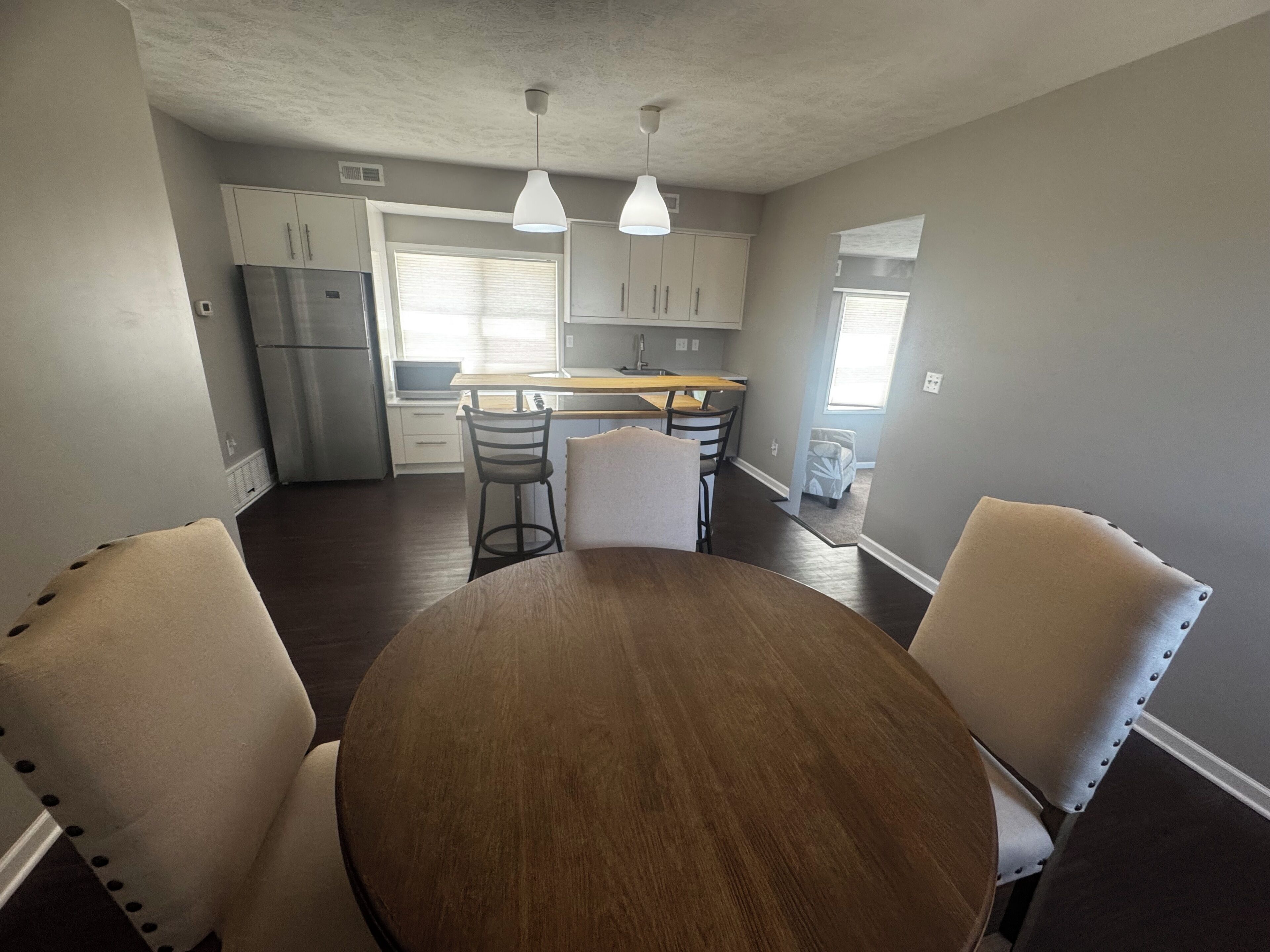 A kitchen with white cabinets, a stainless steel refrigerator, a dining table with four chairs, and pendant lights.