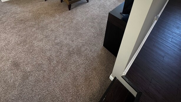 Un salon avec un canapé beige, un pouf assorti, un petit fauteuil et un téléviseur posé sur un meuble.