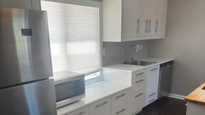 Une cuisine moderne dotée d’appareils électroménagers en acier inoxydable, de meubles blancs, d’un four encastré et d’une fenêtre avec des stores.
