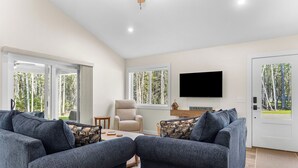 A modern living room with a sofa set, a chair, a coffee table, a flat-screen TV, and large windows with a view of trees. - Seal Point Escape - NEW! (Lamoine)