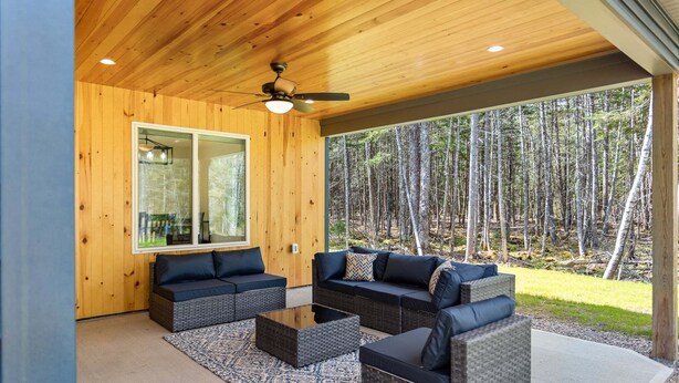 A covered outdoor seating area with a ceiling fan, a large window, and a view of a forest. - Seal Point Escape - NEW! (Lamoine)