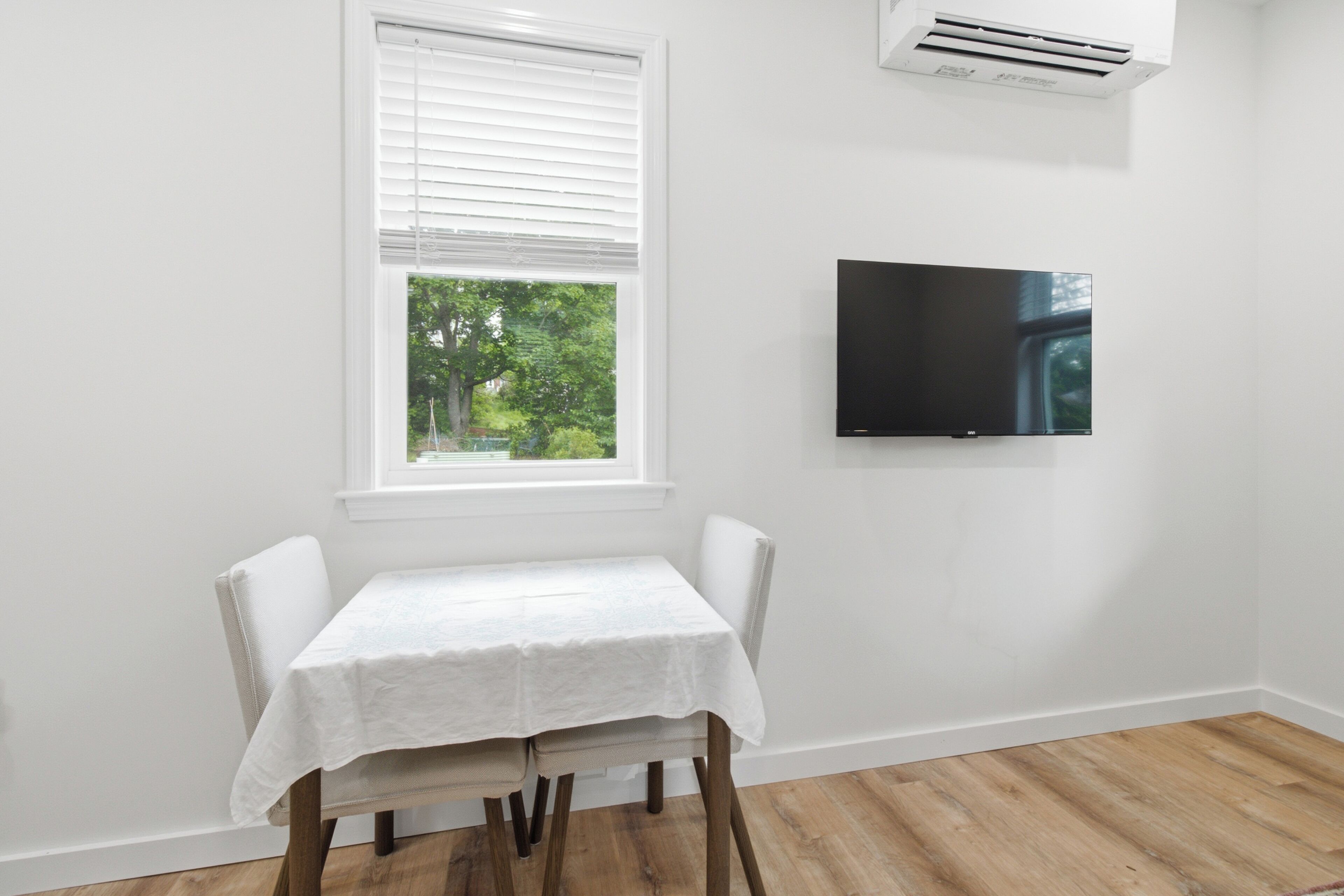 Quarto com uma mesa branca, janela com persianas, televisão na parede e ar condicionado.