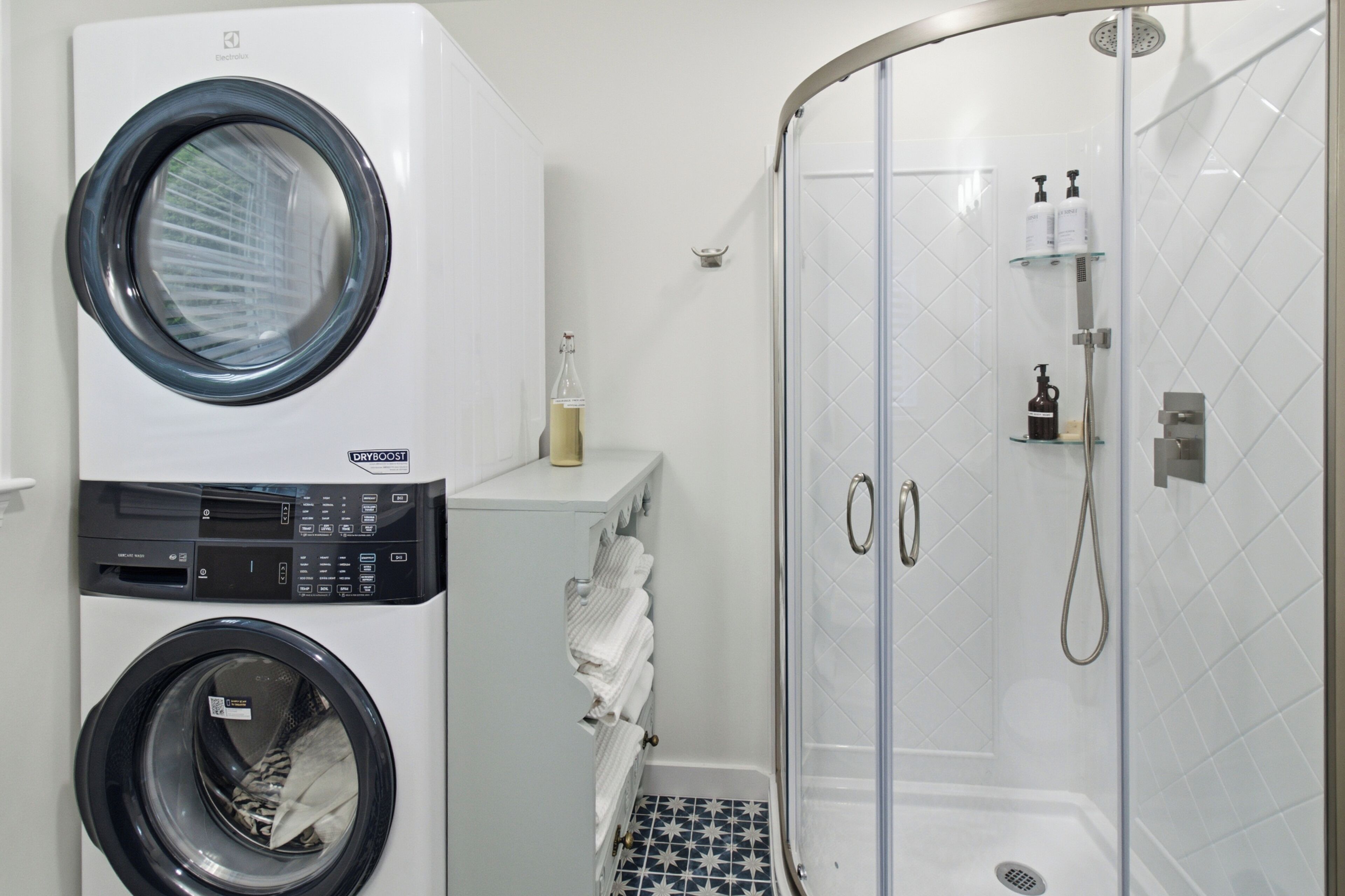A compact laundry area with a front-loading washer and dryer, a glass-enclosed shower, and a small shelf with toiletries.