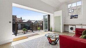 Ein geräumiger Wohnbereich mit einem roten Sofa, einem Glastisch, einer Waschmaschine und Blick durch eine Glastür auf einen Balkon.