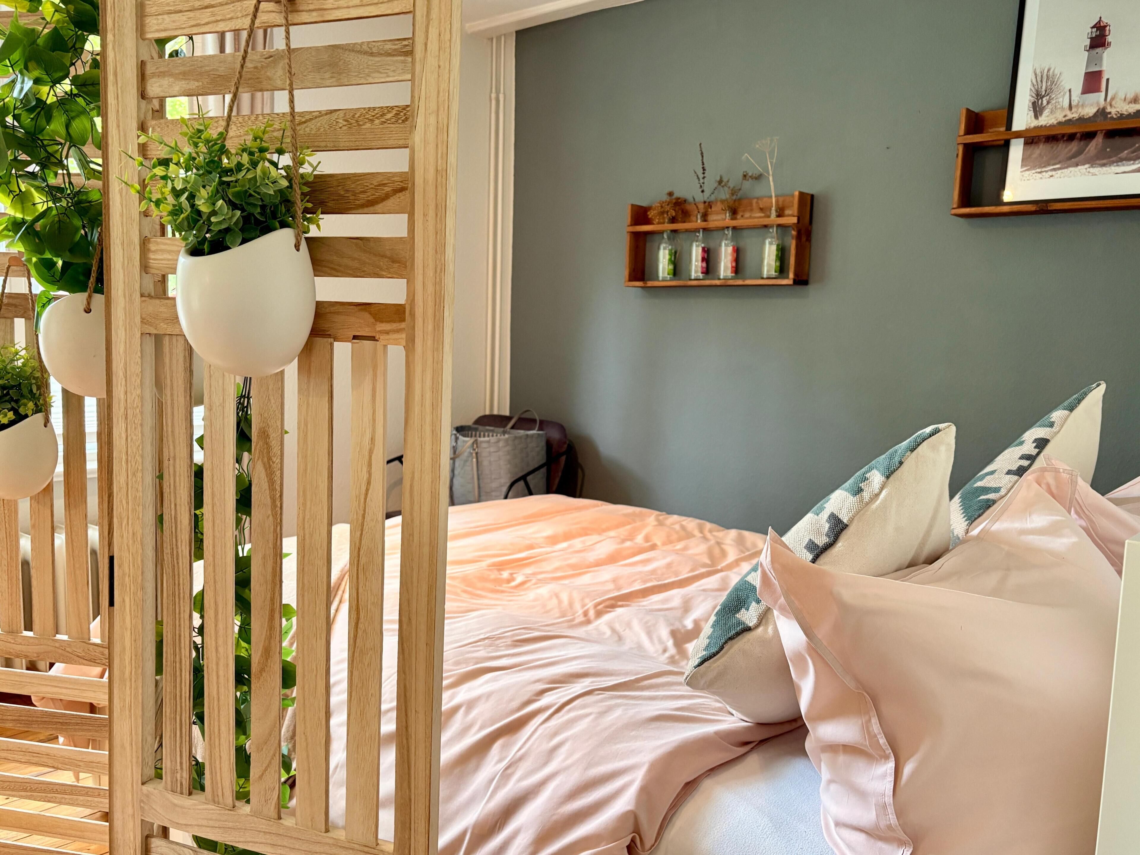 A bedroom with a bed, pillows, a wooden headboard, a wall-mounted shelf with decorative items, and hanging planters.