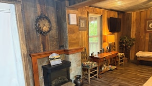 Ein rustikales Interieur mit Kamin, Holzwänden, einer Uhr, einem Willkommensschild, einem Fernseher und einer Bank.