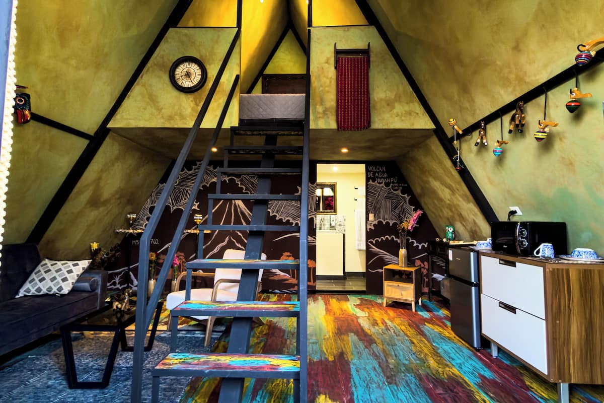 A cozy attic bedroom with a sloped ceiling, a wooden staircase, a bed, a small kitchenette, and a colorful wooden floor.
