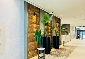 A modern lobby with a herringbone-patterned wall, large potted plants, a marble reception desk, and a tiled floor.