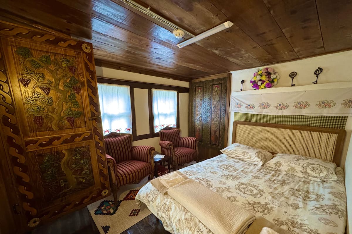 A room with a large bed, two upholstered chairs, a small table, and a decorative wooden panel.
