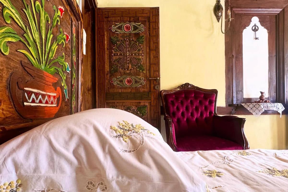 A room with a bed, a chair, a door with intricate designs, and a window with a view.