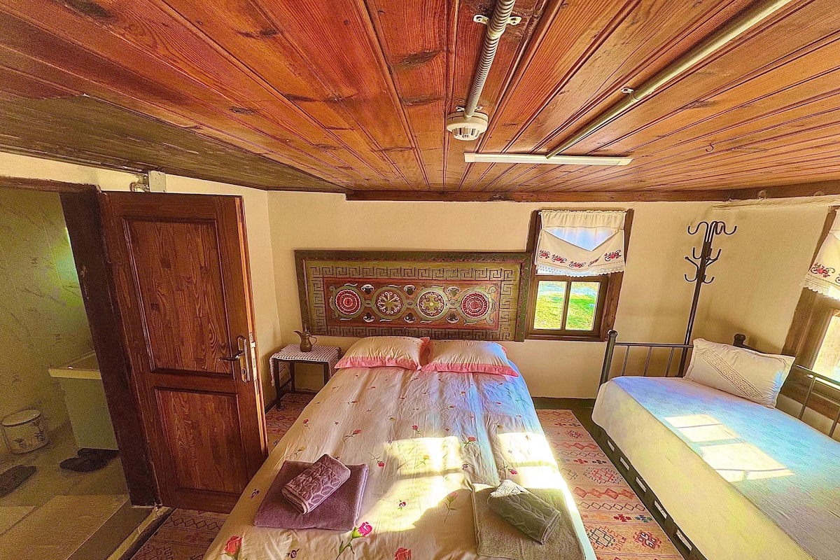 A bedroom with a wooden ceiling, a bed with floral bedding, a bedside table, and a window with a view.