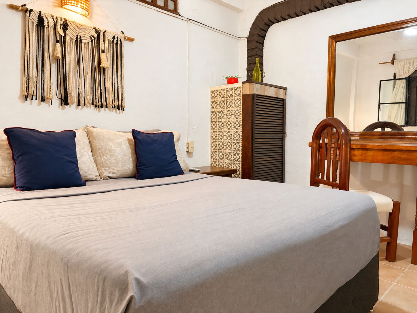 A neatly made bed with a grey bedspread and two pillows, a wall-mounted macrame decoration, a small table with a bottle and a plant, and a wooden chair.