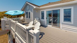 A deck with white chairs and a blue umbrella, a house with large windows, and a clear sky. - Corolla Retreat w/ Views, Hot Tub & Amenities (Corolla)
