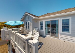 Eine Terrasse mit weißen Stühlen und einem blauen Sonnenschirm, ein Haus mit großen Fenstern und ein klarer Himmel.
