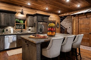 A rustic kitchen with wooden cabinets, a stainless steel refrigerator, and a fruit bowl on the island. - Family Retreat on Fightingtown Creek • HotTub • FirePit • Games • Creek Access (Blue Ridge)