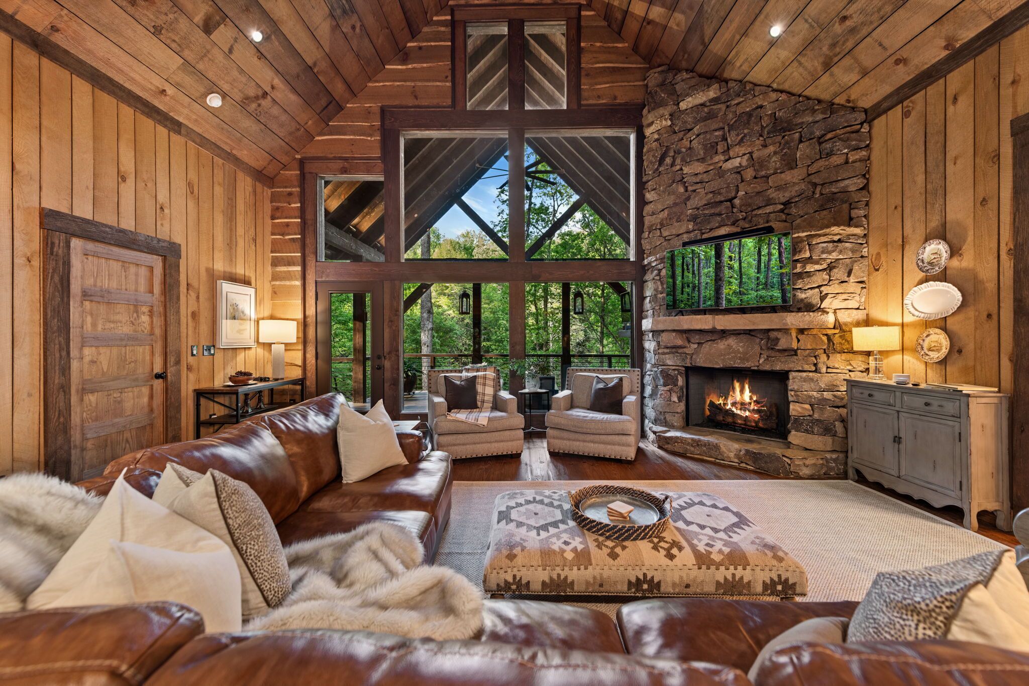 A rustic interior with a stone fireplace, leather sofas, and a large window overlooking greenery.