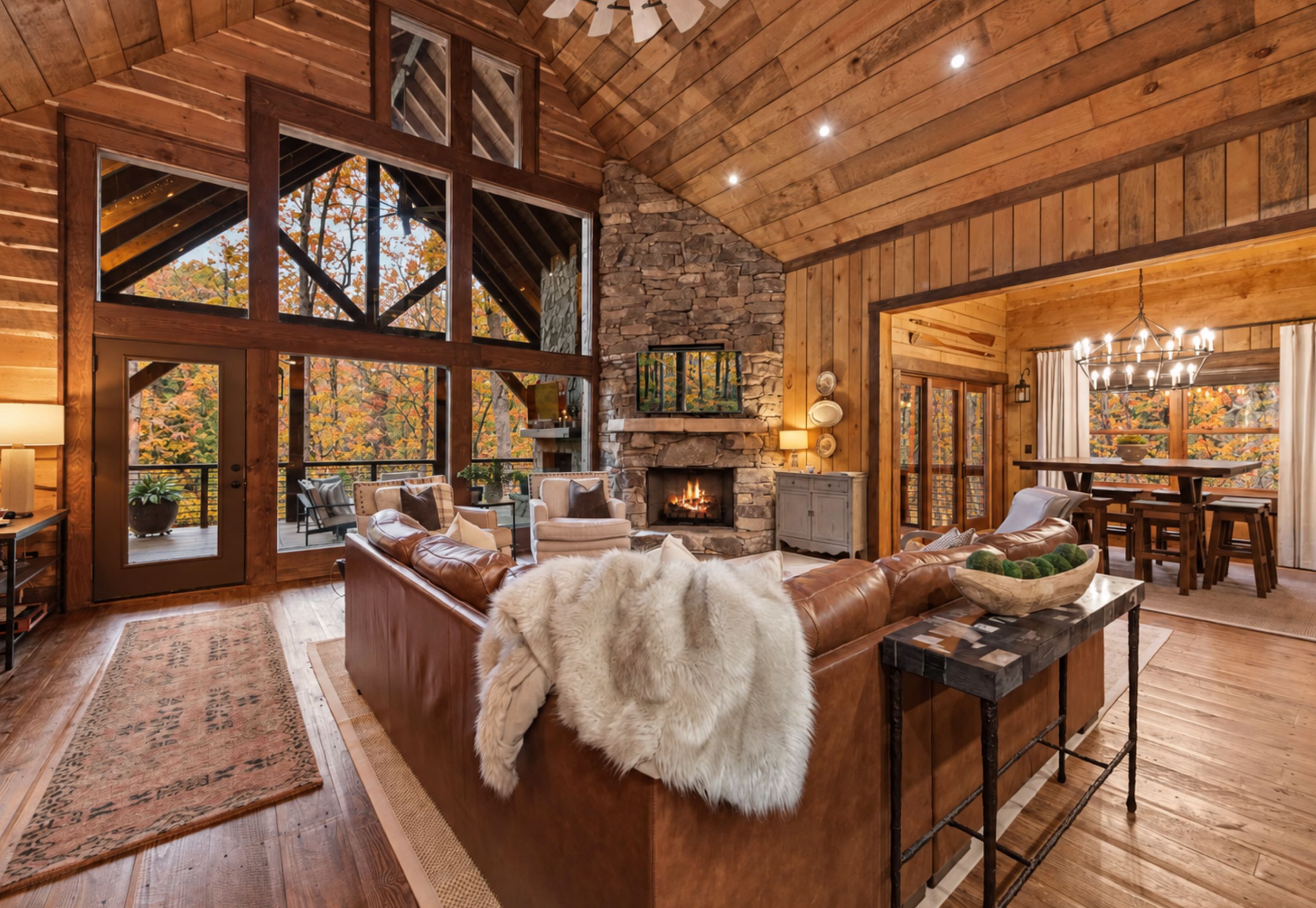 A spacious living area with a stone fireplace, leather furniture, and large windows offering a view of autumn foliage.