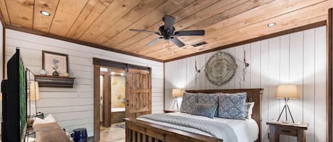 A bedroom with a wooden ceiling, a bed with patterned pillows, a ceiling fan, and a wall-mounted decorative plate.