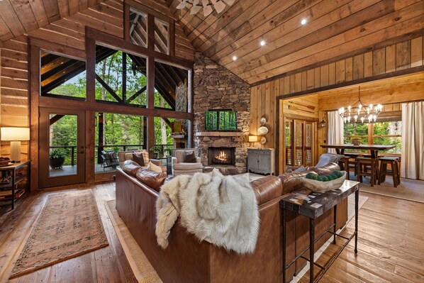 A rustic cabin interior with a stone fireplace, leather seating, and large windows. - Family Retreat on Fightingtown Creek • HotTub • FirePit • Games • Creek Access (Blue Ridge)